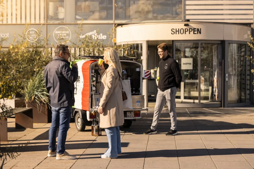 Besøgende foran indgangen til Shoppen i Aalborg nyder kaffe ved en kaffevogn i solskinnet.