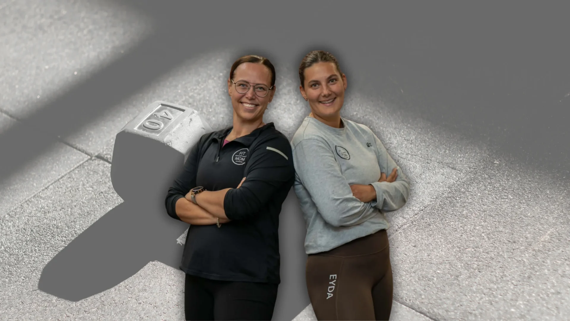 To smilende kvindelige instruktører hos FitMom Aalborg foran en håndvægt i træningscentret.